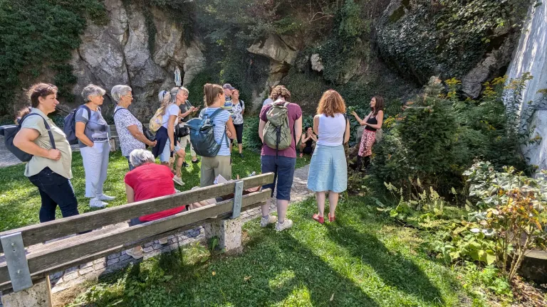 Eine Gruppe von Menschen, darunter Frauen, steht in einem Garten, eine Frau spricht. Eine Frau in einem roten Kleid sitzt auf einer Bank.