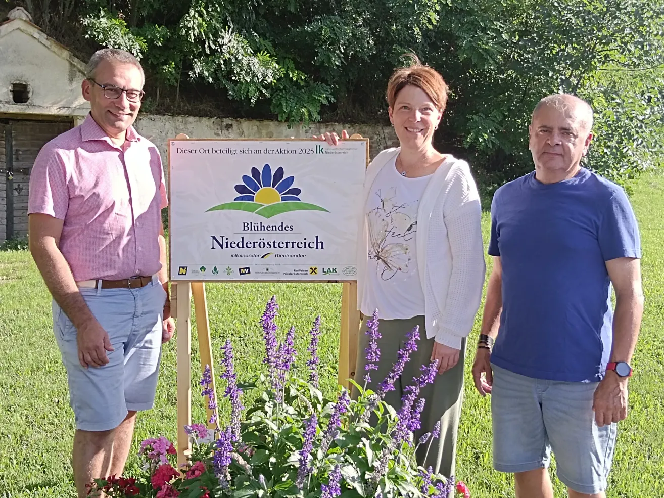 Drei Personen stehen in einem Garten mit einem Schild, das 'Bluhende Niederosterreich' und eine blaue Blume zeigt. Sie lächeln und scheinen für ein Foto zu posieren.
