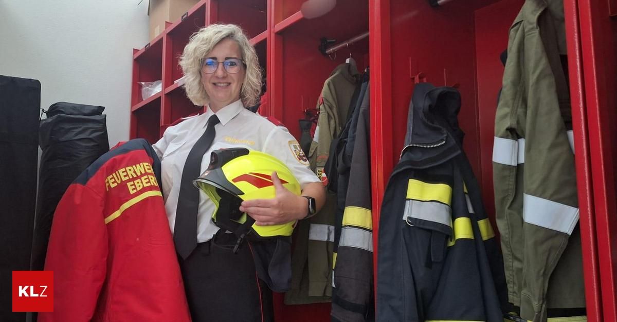 Eine lächelnde Frau in einer Feuerwehruniform, die einen gelben Helm hält, steht in einem Umkleideraum mit roten Regalen, trägt eine Brille und eine Uhr.