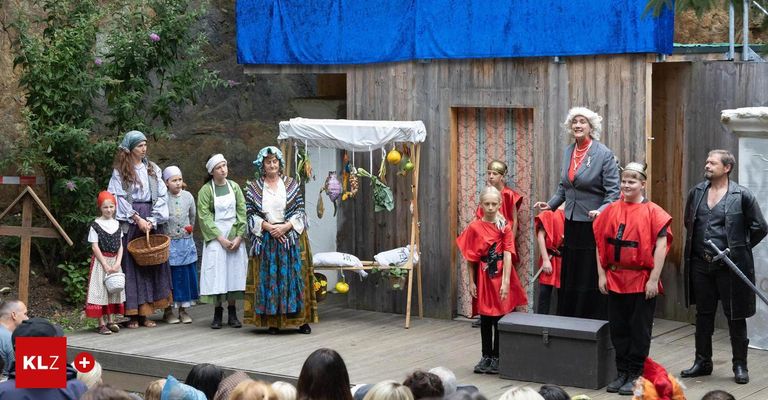 Eine Gruppe von Erwachsenen und Kindern führt ein Theaterstück auf der Bühne auf, wobei eine Erwachsene ein graues Kostüm mit weißer Perücke trägt und ein großer hölzerner Kasten vor ihnen steht.
