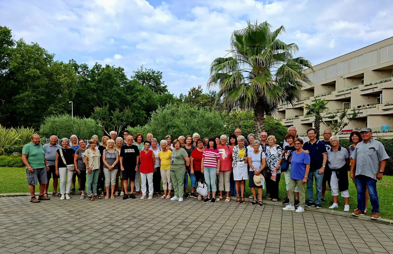 Eine Gruppe von Menschen, lässig gekleidet, posiert für ein Foto in einem Park mit einer Palme und einem Gebäude im Hintergrund.