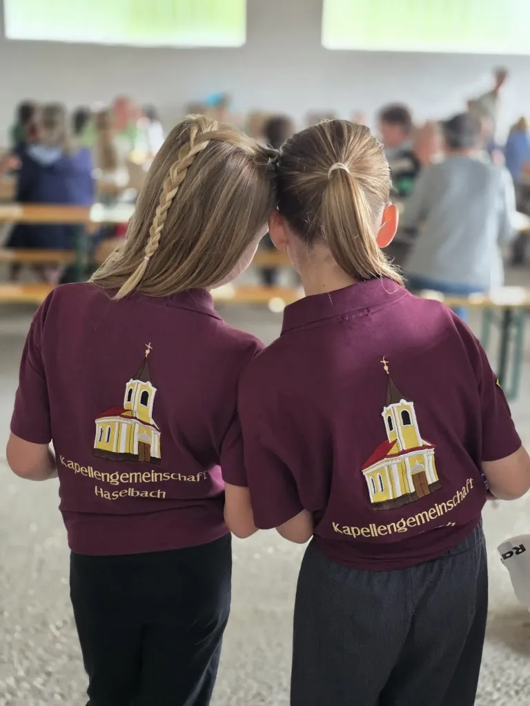 Zwei Frauen mit blonden Haaren in Zöpfen und lilafarbenen Hemden mit einer Kirchengrafik auf dem Rücken stehen in einer Cafeteria.