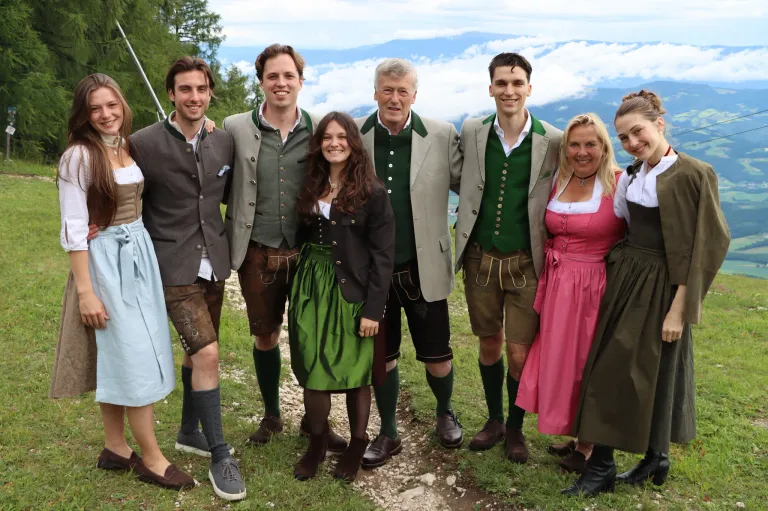 Eine Gruppe von Menschen in traditioneller deutscher Kleidung, die lächelnd und posierend für ein Foto auf einem Grasfeld mit Bergen im Hintergrund stehen.