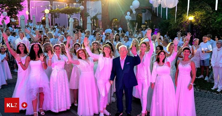 Eine Gruppe von Menschen in rosa Kleidern und weißen Hüten versammelt sich draußen. Ein Mann im Anzug winkt der Menge zu. Im Hintergrund befinden sich Luftballons und Lichter.