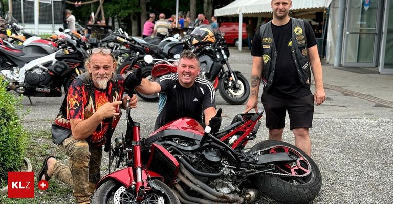 Drei Männer stehen neben einer umgestürzten roten Motorräder. Der Mann auf der linken Seite trägt ein Flammen-Muster-Shirt und einen Bart. Der Mann in der Mitte trägt ein schwarzes T-Shirt mit einem Streifen. Der Mann auf der rechten Seite trägt eine schwarze Weste. Hinter ihnen sind Motorräder geparkt und Menschen versammelt.
