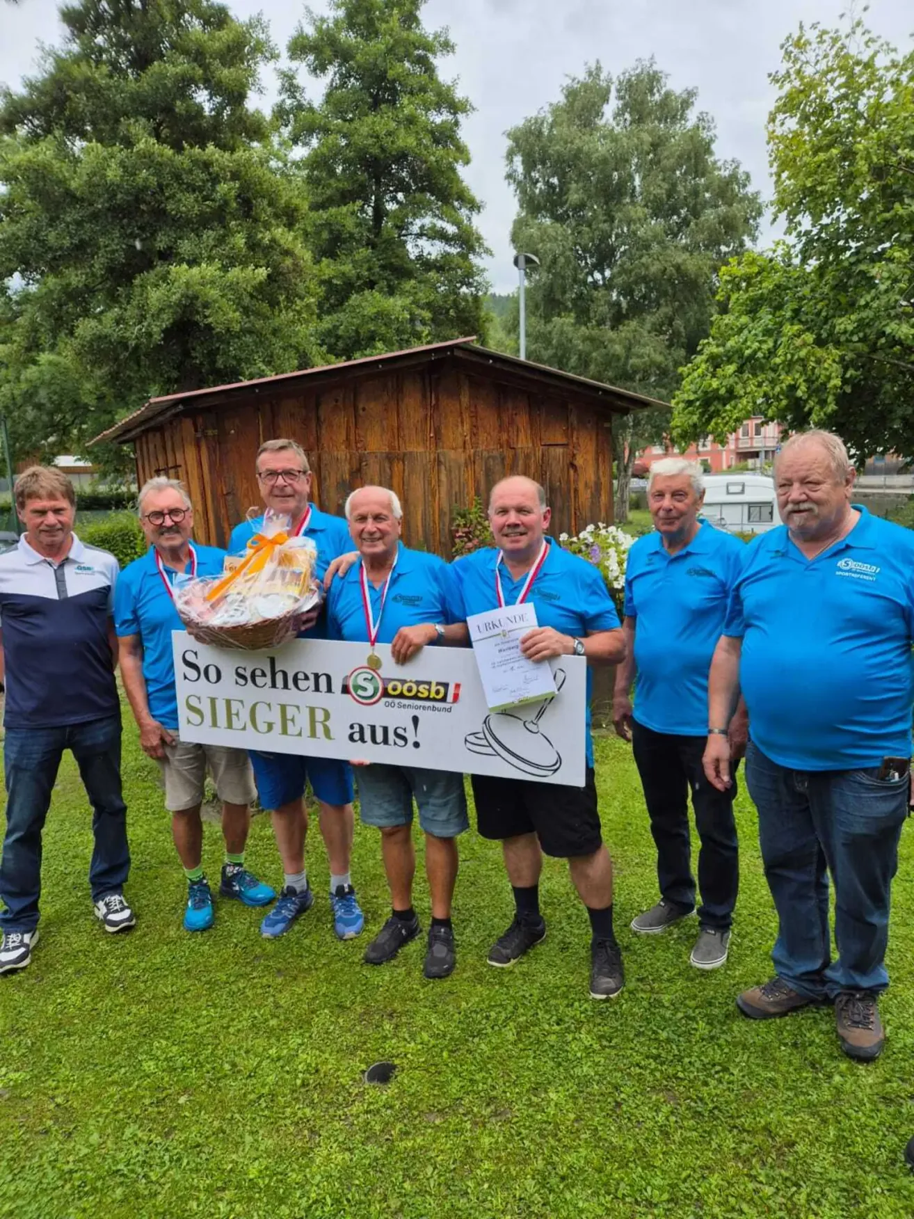 Eine Gruppe von Männern in blauen Hemden und Medaillen posiert mit einem Korb und einem Schild, das 'So sehen Sieger aus!' in einem Rasenbereich in der Nähe eines Holzhauses liest.