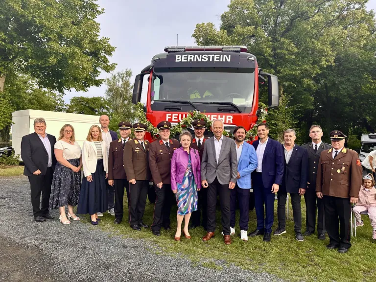 Eine Gruppe von Menschen, darunter Feuerwehrleute, steht vor einem roten Feuerwehrwagen mit 'Bernstein' und 'Feuerwehr' darauf geschrieben. Sie tragen alle formelle Kleidung.