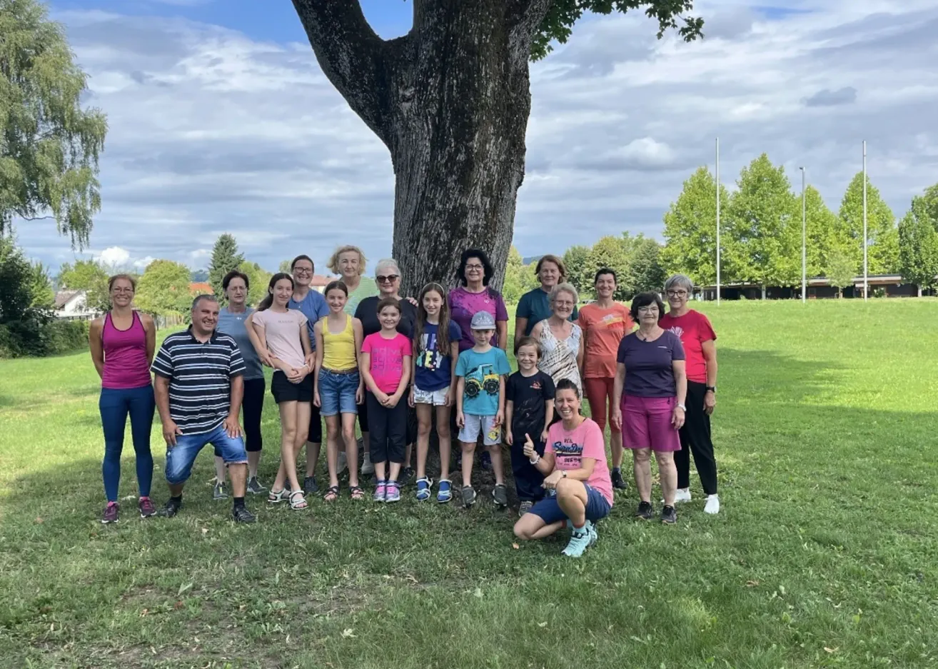 Eine Gruppe von Menschen posiert für ein Foto in einem Park mit einem Baum in der Mitte. Es gibt viele Menschen in verschiedenen Kleidungsstücken. Sie alle lächeln.