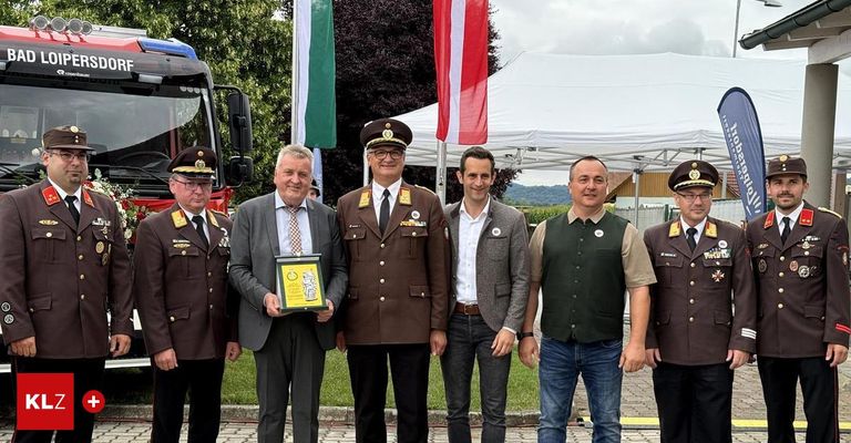 Sechs Männer in Uniformen stehen vor einem Zelt mit Flaggen und lächeln. Einer hält eine Auszeichnung. Dahinter steht ein LKW und grüne Bäume.