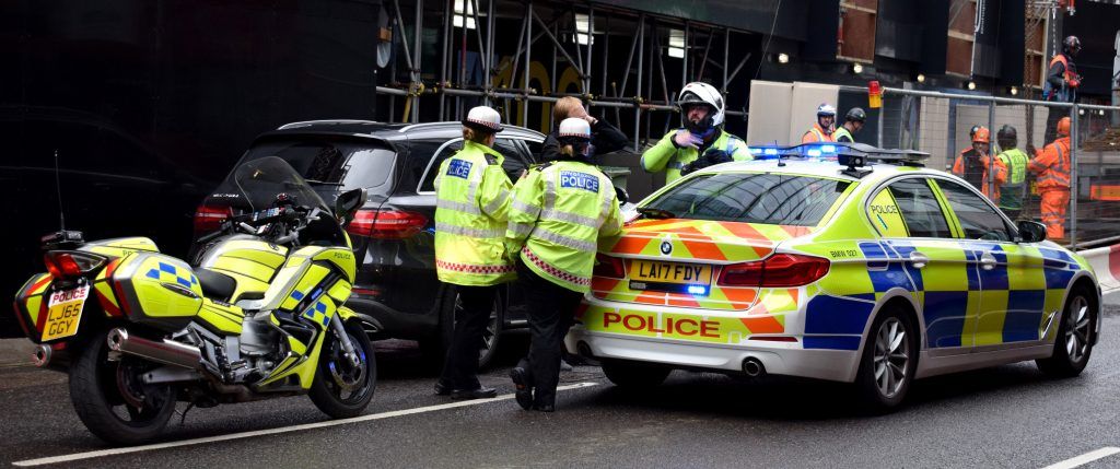 Polizisten in Warnwesten umgeben ein schwarzes Auto. Ein Polizeimotorrad ist in der Nähe geparkt. Die Szene befindet sich in der Nähe einer Baustelle.