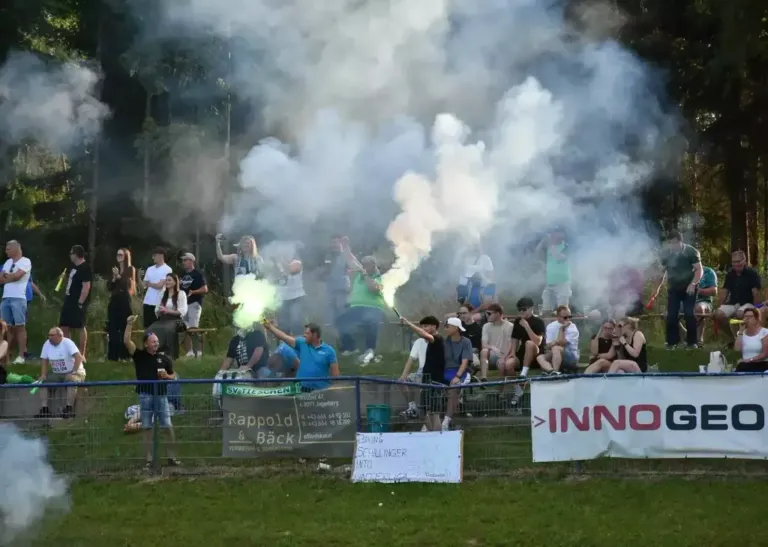 Eine Menschenmenge bei einer Outdoor-Veranstaltung, einige halten Rauchbomben und erzeugen einen rauchigen Effekt. Ein Mann hält eine grüne Rauchbombe und ein anderer eine weiße. Banner sind am Zaun zu sehen, und einige Leute sitzen auf dem Boden.