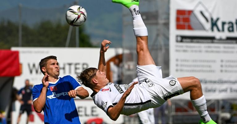 Ein Fußballspieler in Weiß springt, um einen Ball zu köpfen, während ein anderer Spieler in Blau versucht, zu blocken.