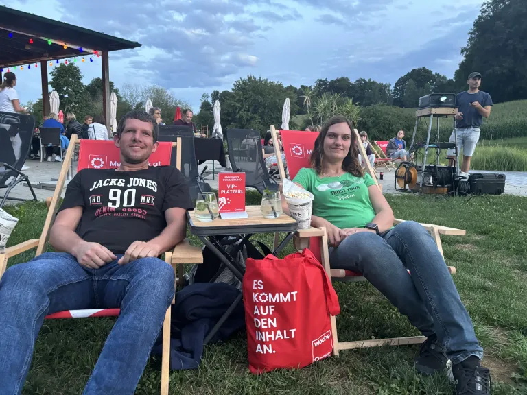 Zwei Personen sitzen in Klappstühlen draußen mit Getränken und Popcorn. Der Mann trägt ein Jack & Jones-Shirt. Die Frau trägt ein grünes Shirt mit Trust Your Intuition. Eine rote Tasche mit Es Kommt Auf Den Inhalt An. steht auf dem Gras.