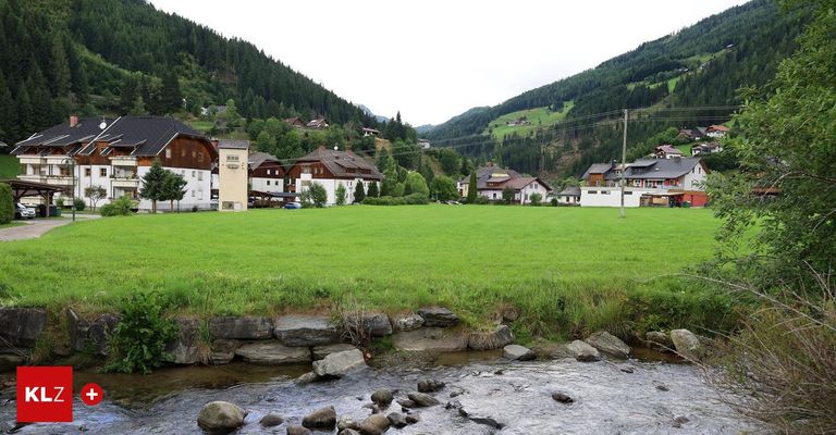 Eine ländliche Szene mit einem Bach, Häusern und Bergen im Hintergrund. Die Häuser sind von üppigen grünen Feldern und Bäumen umgeben.