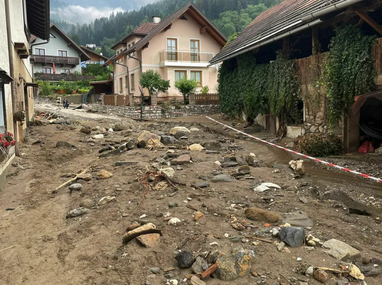Ein schlammiger Bereich mit verstreuten Steinen und Schutt, ein Haus mit Balkon und roter Fahne und ein Gebäude mit grünen Ranken.