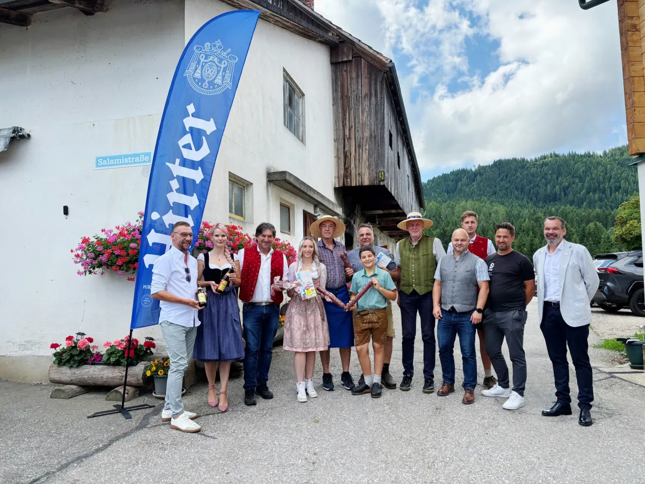 Eine Gruppe von Menschen in traditioneller Kleidung posiert für ein Foto vor einem Gebäude, mit Bergen im Hintergrund. Eine blaue Fahne mit weißem Text ist links sichtbar.