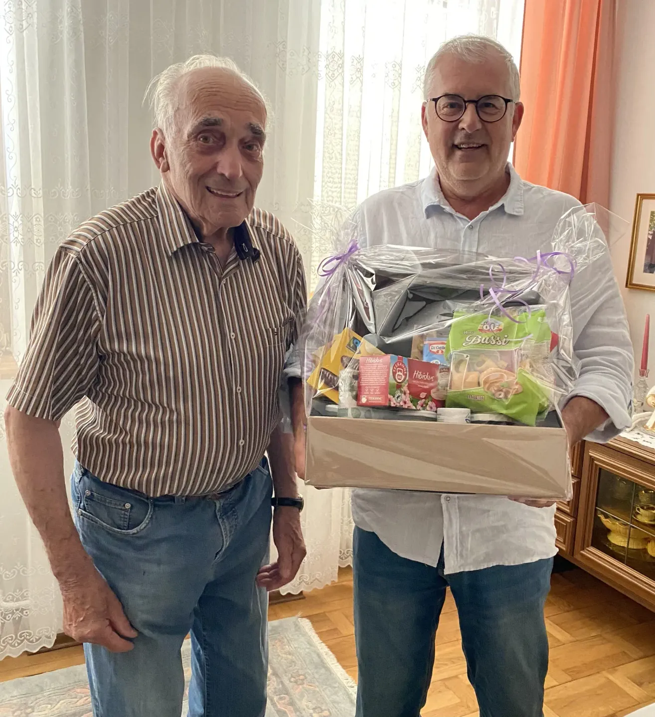 Zwei Männer stehen nebeneinander und halten eine Schachtel mit Essen vor einem Fenster.