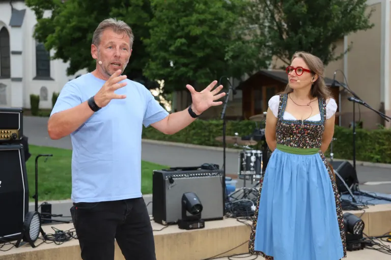 Ein Mann und eine Frau stehen in einem Außenbereich, wahrscheinlich für eine Aufführung. Der Mann spricht und gestikuliert mit den Händen. Die Frau trägt ein traditionelles bayerisches Kleid. Hinter ihnen stehen Musikinstrumente und Ausrüstung. In der Ferne befinden sich Bäume und Gebäude.