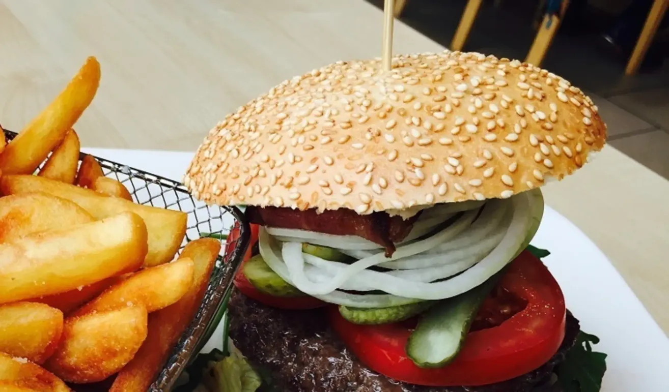 Nahaufnahme eines Burgers mit Sesamkörnern auf dem Brötchen, mit Rinderpattie, Speck, Tomate, Zwiebeln und Gurken. Daneben liegen Pommes Frites.