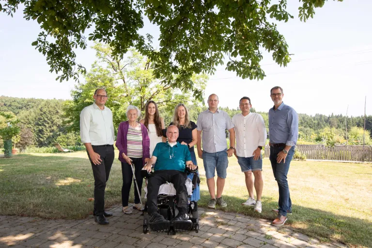 Sieben Menschen, darunter ein Mann im Rollstuhl, stehen auf einem gepflasterten Weg in einem Rasenbereich. Hinter ihnen spendet ein Baum Schatten. Der Mann im Rollstuhl lächelt, und die anderen stehen hinter ihm.