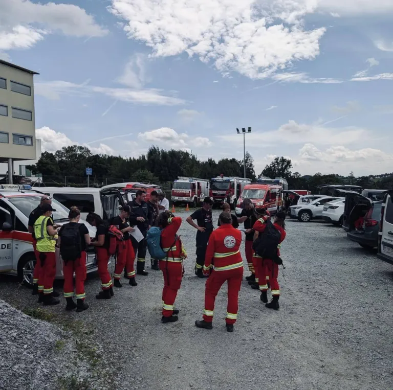 Eine Gruppe von Menschen in roten Uniformen versammelt sich auf einem Parkplatz, möglicherweise um etwas zu besprechen. Einige von ihnen tragen Rucksäcke. In der Ferne befinden sich Autos, ein Gebäude und Bäume.