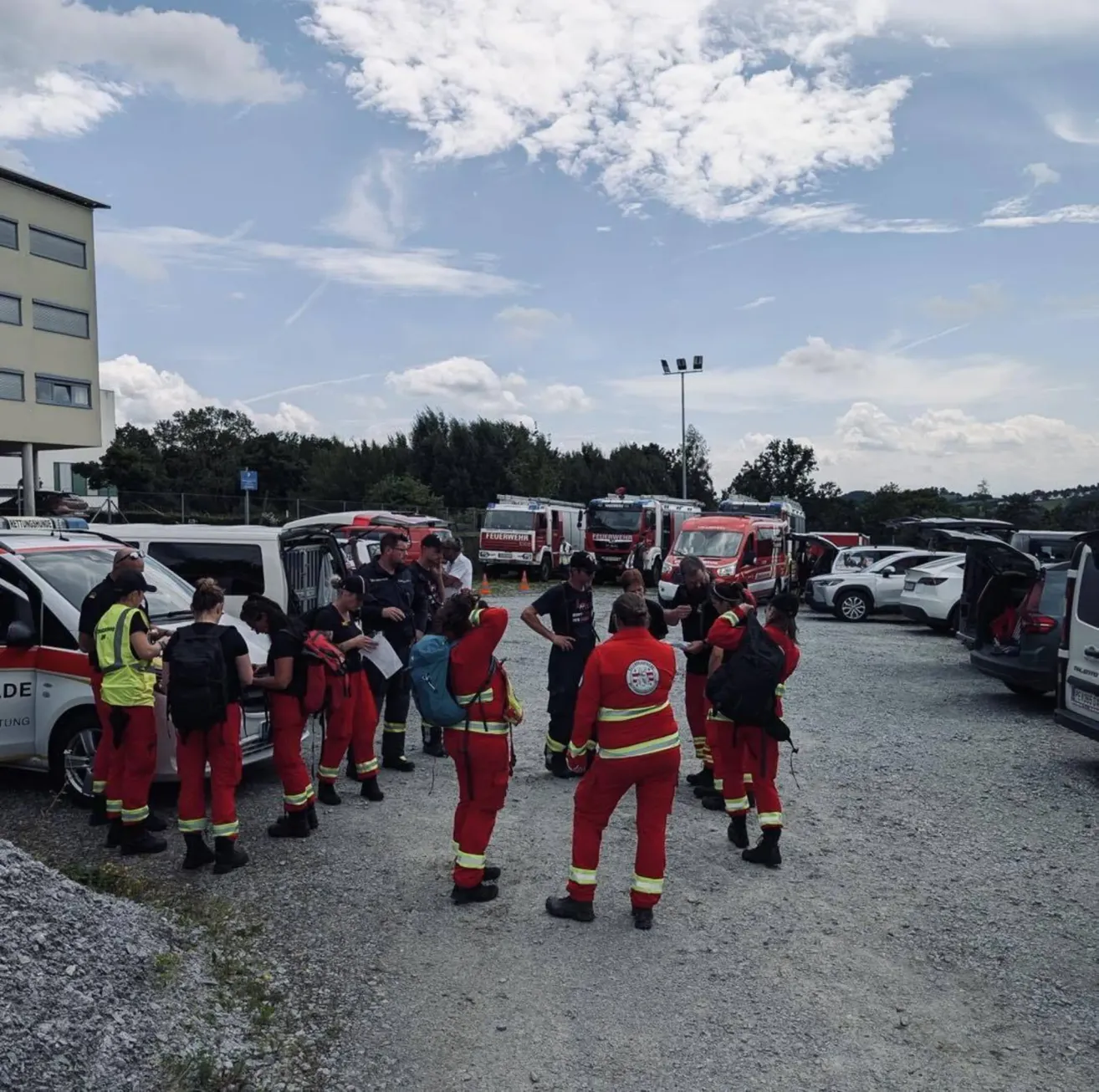 Eine Gruppe von Menschen in roten Uniformen versammelt sich auf einem Parkplatz, möglicherweise um etwas zu besprechen. Einige von ihnen tragen Rucksäcke. In der Ferne befinden sich Autos, ein Gebäude und Bäume.