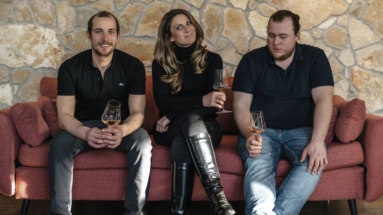 Drei Personen sitzen auf einer roten Couch und lächeln. Zwei Männer halten Weingläser und eine Frau in der Mitte hält auch eines. Sie scheinen für ein Foto vor einer Steinmauer zu posieren.