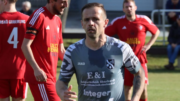 Drei Männer stehen auf einem Grasfeld und tragen Sporttrikots. Der Mann in der Mitte trägt ein graues Shirt mit 'E&I Integer' und 'Elektrotechnik Eberhardt' darauf. Die Männer hinter ihm tragen rote Trikots mit den Worten 'Kalc BENN' und 'Kalc' darauf.