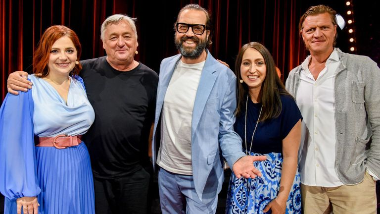 Drei Personen posieren für ein Foto. Ein Mann in einem blauen Blazer, eine Frau in einem blauen Kleid und ein Mann in einem schwarzen T-Shirt. Hinter ihnen ein roter Vorhang.