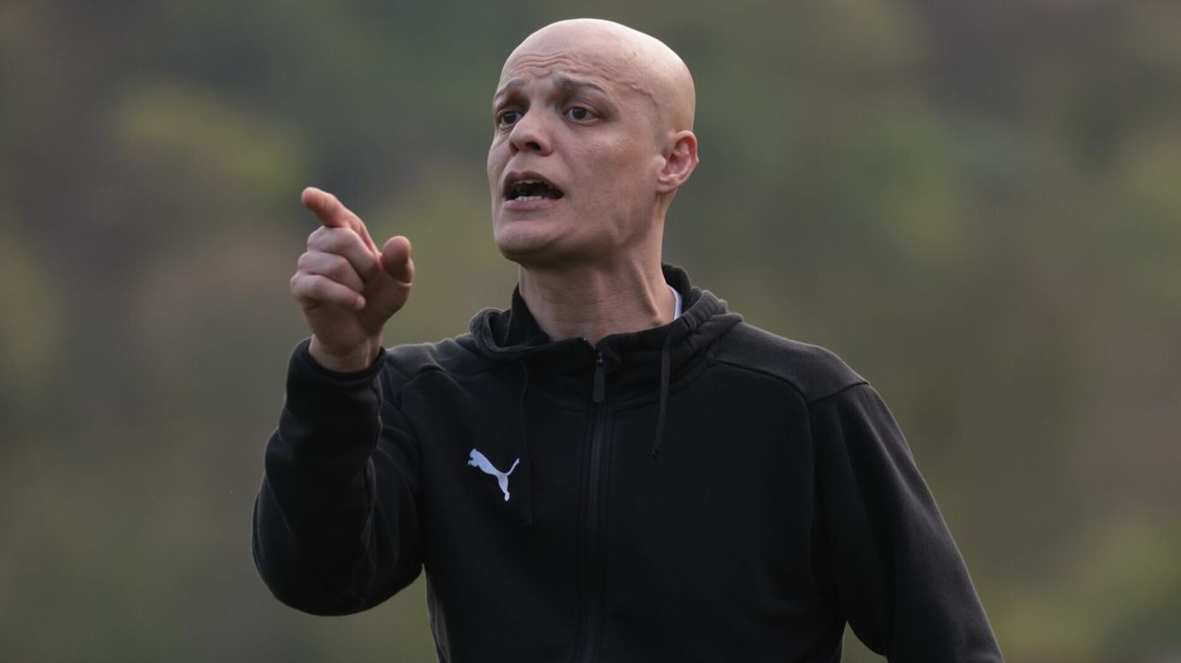Ein kahlköpfiger Mann in einem schwarzen Puma-Hoodie spricht und zeigt zur Seite, möglicherweise in einer sportlichen Trainingsumgebung.