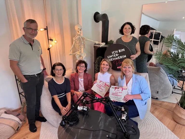 Mehrere Frauen und ein Mann posieren mit einem Skelett und einem Schild in einem Raum. Die Frau in der Mitte hält einen Beutel mit dem Text 'ICH BIN UNTER-NEHMERIN'.