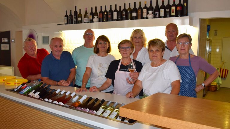 Eine Gruppe von Menschen, wahrscheinlich Angestellte, steht hinter einer Bar mit Regalen voller Weinflaschen darüber. Einige halten Weingläser und tragen Brillen.