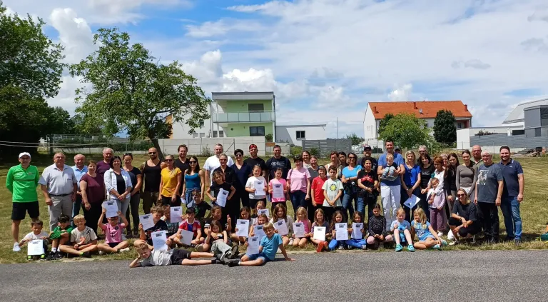 Eine Gruppe von Erwachsenen und Kindern posiert für ein Foto im Freien vor einigen Häusern. Alle halten ein Zertifikat.