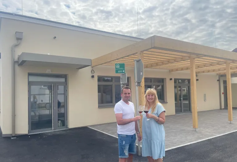Zwei Personen stehen vor einem Gebäude und halten Weingläser in der Hand. Ein Mann trägt ein weißes T-Shirt und Jeansshorts, während eine Frau ein blaues Kleid trägt. Beide lächeln. Hinter ihnen befindet sich ein Schild mit der Nummer B311.