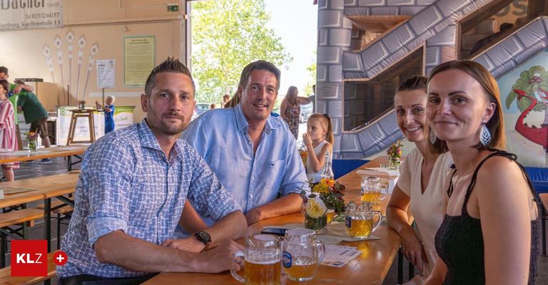 Vier Personen sitzen an einem Tisch mit Bierkrügen und Blumen davor. Hinter ihnen steht eine Frau neben einem Kind, und eine aufblasbare Rutsche befindet sich an der Wand.