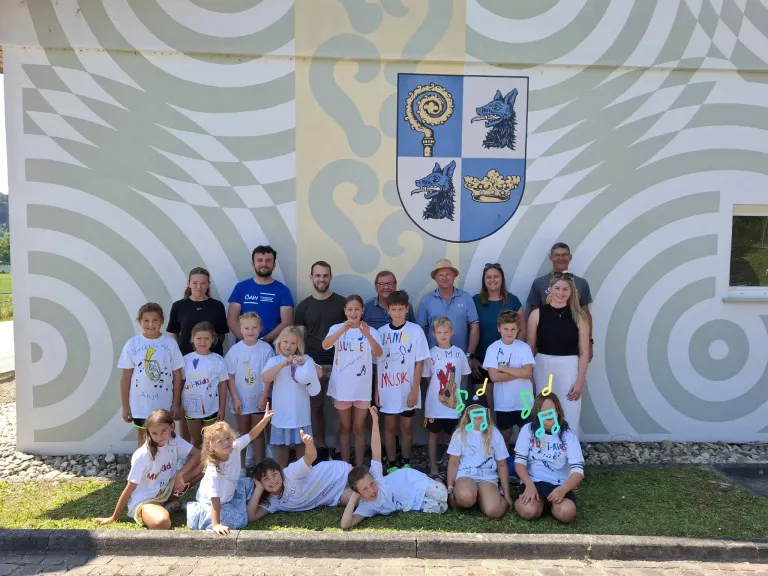 Eine Gruppe von Kindern und Erwachsenen posiert für ein Foto vor einer Wand mit einem Wappen. Die Kinder tragen weiße T-Shirts mit Zeichnungen und Noten.