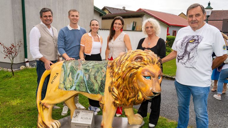 Fünf Personen stehen um eine Löwenstatue und lächeln. Der Löwe hat eine grüne Decke mit dem Wort Roma und einer Karte. Sie stehen auf einem Rasenfeld mit Häusern im Hintergrund.