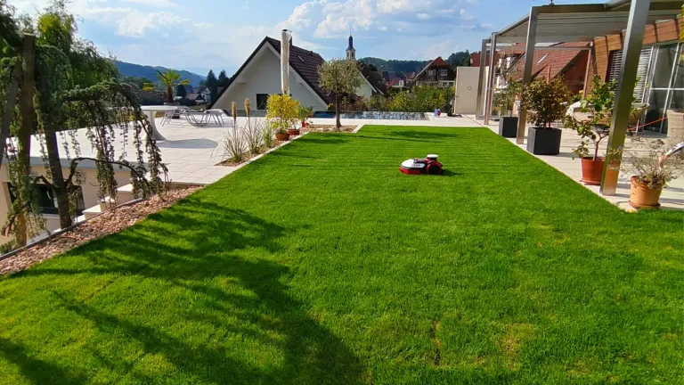 An einem sonnigen Tag zeigt sich ein gepflegter Hinterhof mit einem roten und weißen Roboter-Rasenmäher. Dahinter ist ein modernes Haus mit braunem Dach und einem Pool zu sehen.