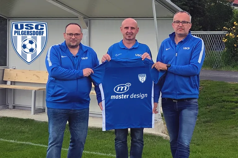 Drei Männer in blauen Jacken stehen auf einem Feld. Einer hält ein blaues Trikot mit dem Logo und dem Text 'master design'. Im Hintergrund ist eine weiße Struktur und ein Logo auf der linken Seite zu sehen.