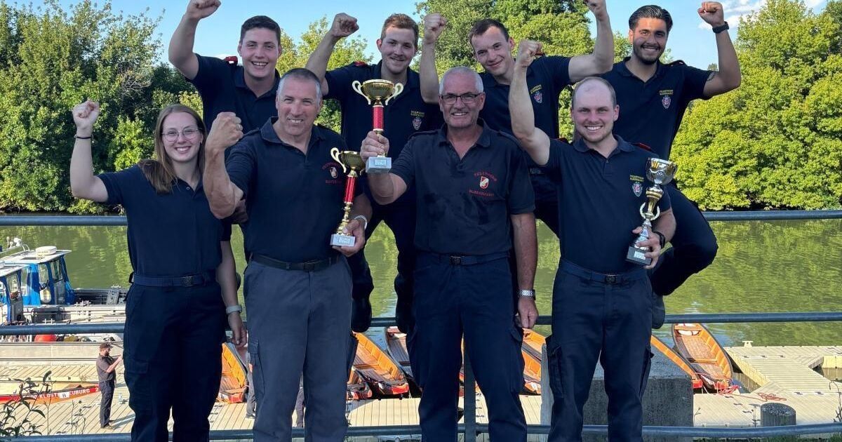 Eine Gruppe von Menschen in dunkelblauen Uniformen mit roten Aufnähern auf den Ärmeln steht auf einem Dock und posiert für ein Foto mit Trophäen. Hinter ihnen sind mehrere kleine Boote verankert, und das Gebiet ist von Bäumen umgeben.