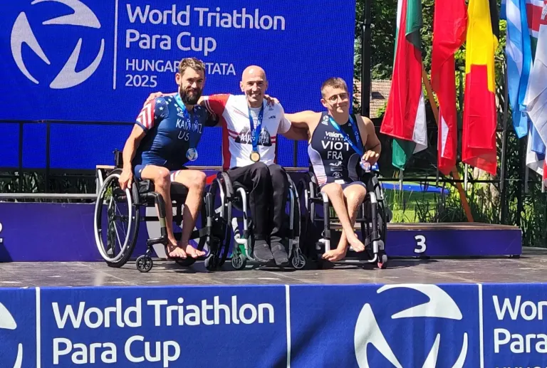 Drei Männer in Rollstühlen posieren für ein Foto auf dem Podium. Hinter ihnen steht ein blauer Banner mit der Aufschrift World Triathlon Para Cup. Der Mann auf der linken Seite trägt eine Medaille um den Hals.