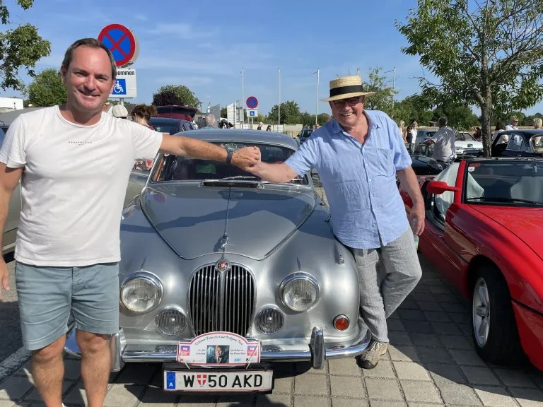 Zwei Männer stehen neben einem silberfarbenen Auto mit dem Kennzeichen W50AKD. Sie lächeln beide und scheinen für ein Foto zu posieren. Das Auto ist auf einem Parkplatz abgestellt.