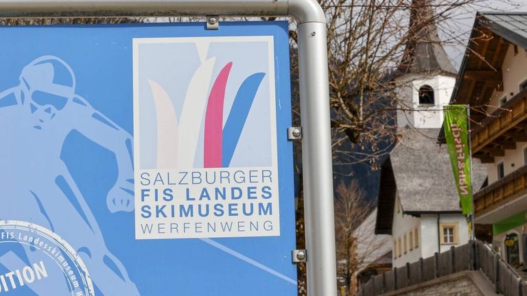 Ein blaues Schild mit weißem Rand und den Worten Salzburger FIS Landes Skimuseum Werfenweng. Der Hintergrund ist blau mit weißen und rosa Linien.
