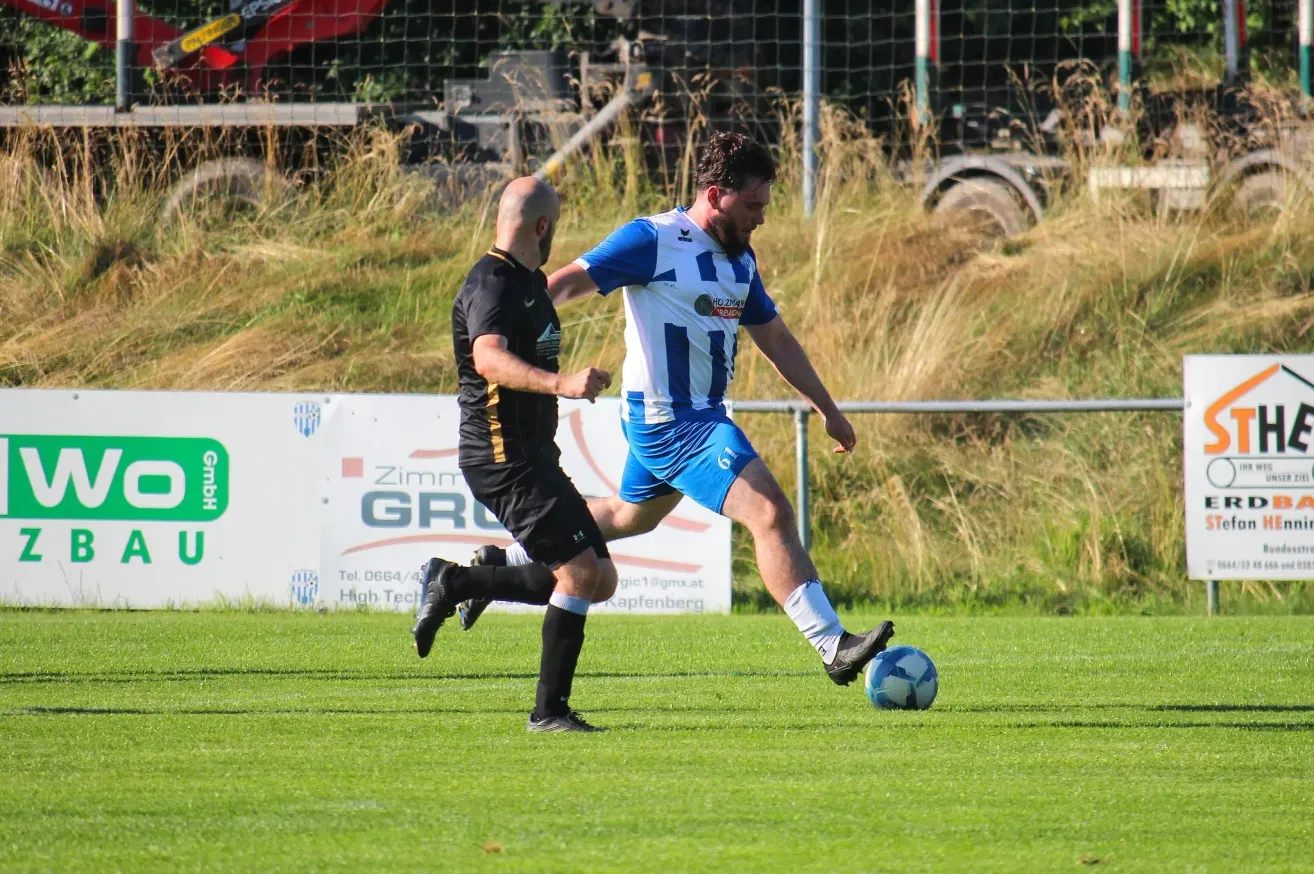Zwei Fußballspieler sind mitten in einer Aktion auf einem Feld, einer in einem blauen Trikot und der andere in Schwarz. Der Spieler in Blau tritt den Ball, während der Spieler in Schwarz versucht, ihn zu blockieren. Dahinter ist ein weißes Banner mit Text.