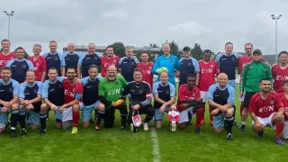 Eine Fußballmannschaft mit Spielern in verschiedenen Farben posiert für ein Foto auf einem Feld. Sie tragen Trikots mit den Buchstaben EVN und halten Pokale. Im Hintergrund sind Bäume und Gebäude zu sehen.