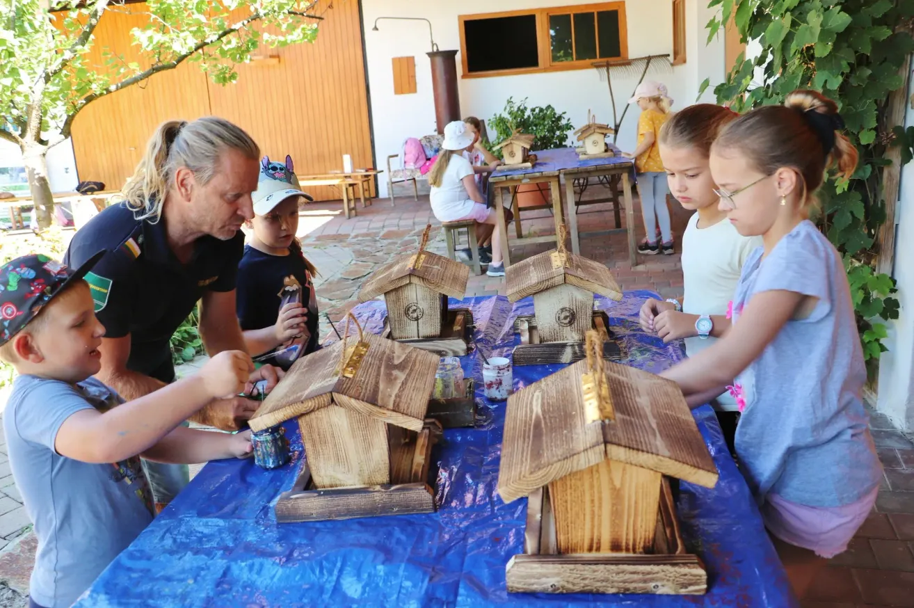 Eine Gruppe von Kindern und einem Erwachsenen malt Holzvogelhäuschen auf einem mit einer blauen Plane bedeckten Tisch. Sie sind draußen in der Nähe eines Hauses mit mehreren Vogelhäuschen.