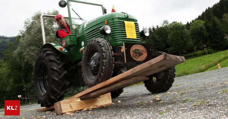 Ein Mann in einem roten Hut fährt einen grünen Traktor und zerquetscht Holz auf einer Kiesstraße. Ein gelbes Papier ist an der Vorderseite des Traktors befestigt.