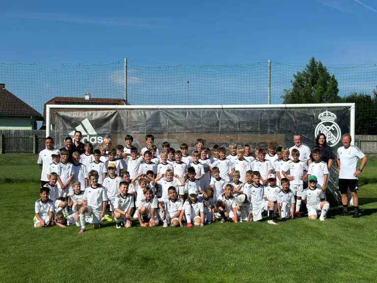 Eine Fußballmannschaft posiert für ein Foto auf einem Rasenfeld, im Hintergrund ein großes Banner. Das Banner trägt das Adidas-Logo und das Real Madrid-Wappen.