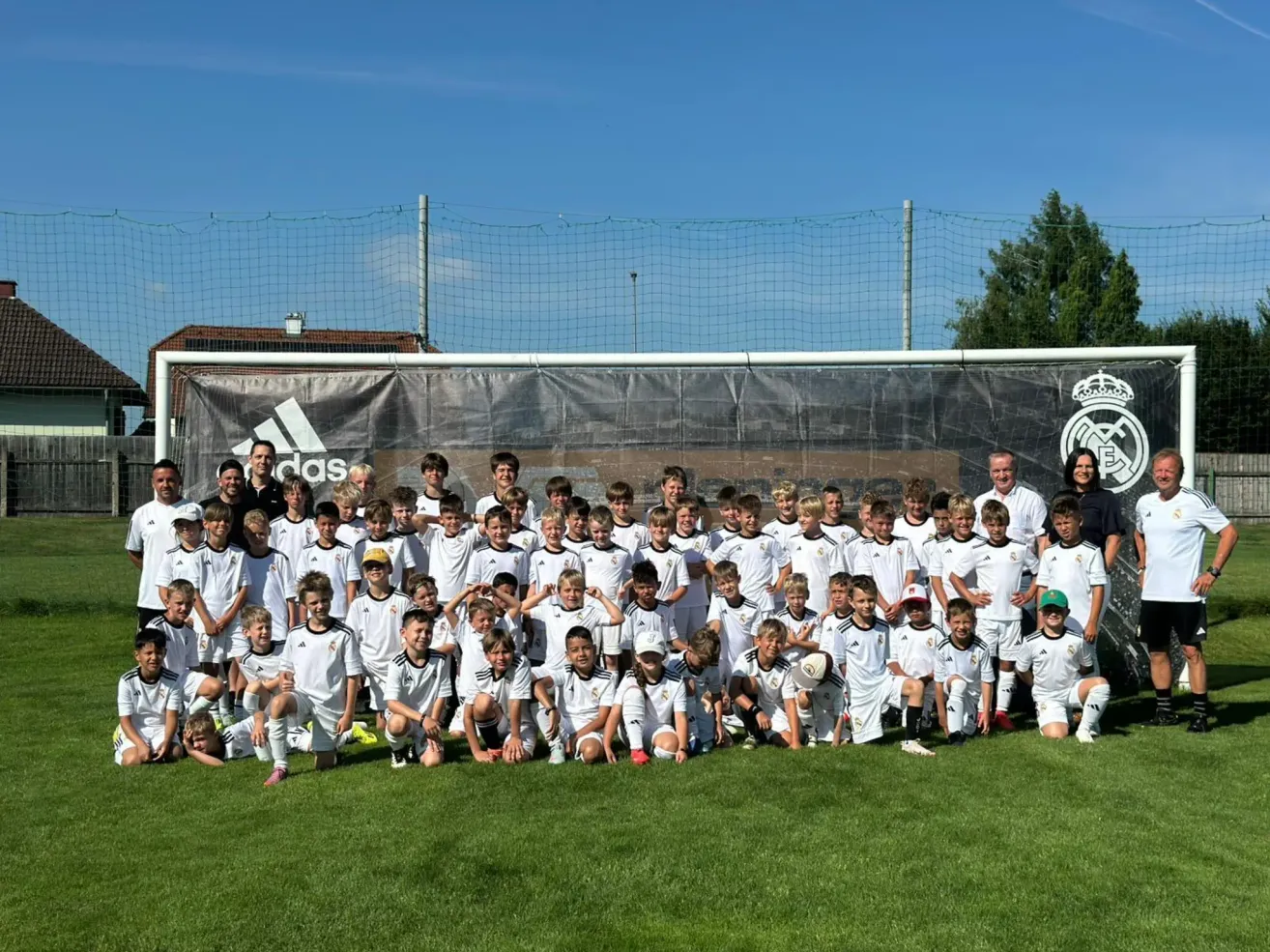 Eine Fußballmannschaft posiert für ein Foto auf einem Rasenfeld, im Hintergrund ein großes Banner. Das Banner trägt das Adidas-Logo und das Real Madrid-Wappen.