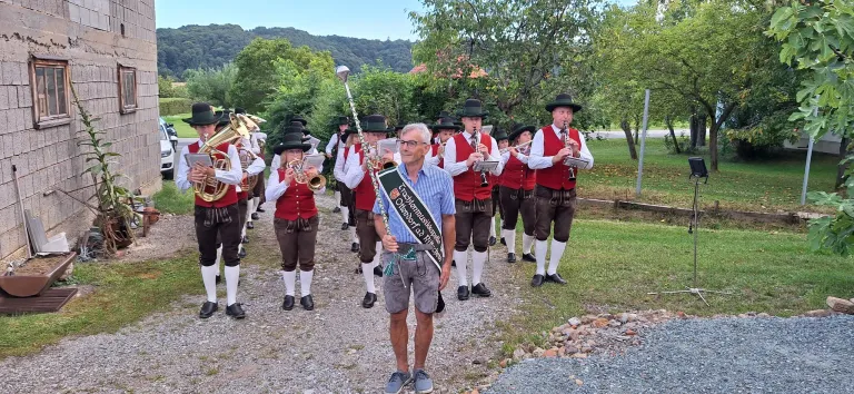 Eine Gruppe von Musikern in traditioneller deutscher Kleidung marschiert in einer Reihe. Sie halten Musikinstrumente in den Händen, darunter Trompeten und Flöten. Ein Mann vorne trägt eine Brille und einen Schal um die Taille. Im Hintergrund sind Bäume und ein Berg zu sehen.